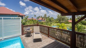 A wooden deck with a pool, lounge chairs, and a view of a tropical landscape with palm trees and a red-roofed building. - Villa Kyneos, prestigious villa on the beach at Cap Est Martinique  (Le François)
