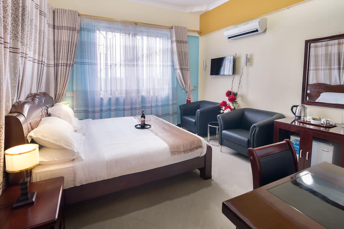 Une chambre d’hôtel comprenant un lit, une table de chevet, une lampe, des rideaux, un téléviseur, un bureau avec une chaise et une petite kitchenette.