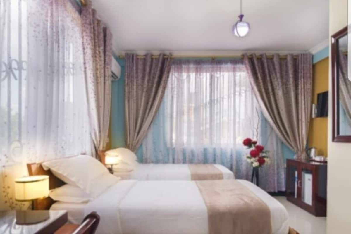Une chambre d’hôtel avec deux lits, une table de chevet avec une lampe, un miroir et une fenêtre avec des rideaux.