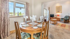 A dining area with a round wooden table set for four, flanked by two chairs with floral upholstery. A television and a small table with a lamp are visible in the background. - The Old Greenhouse - UK45328 (Wragby)