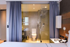 Une chambre d’hôtel moderne dotée d’une douche à parois de verre, de toilettes blanches et d’une table de chevet avec un téléphone et une lampe.