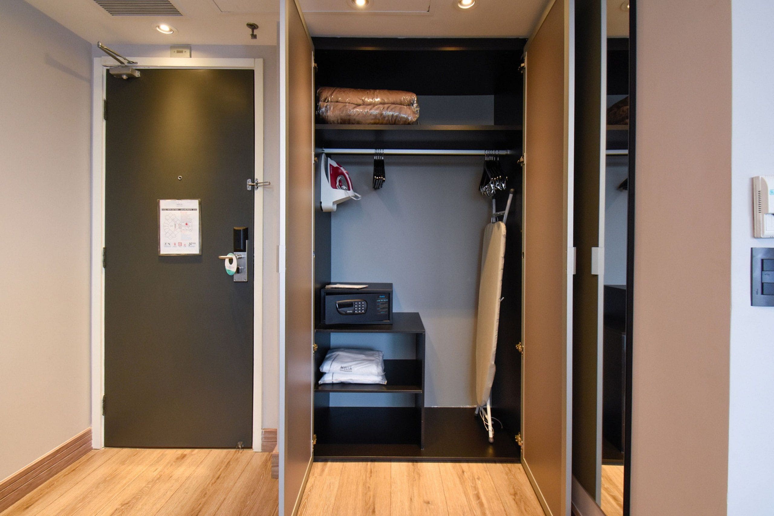 A hotel room with a wooden floor, a dark door, a small shelf with a microwave, and a closet with hanging clothes.