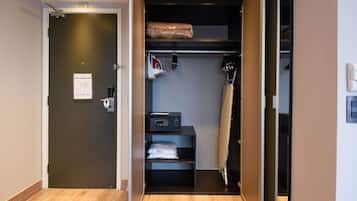 Quarto de hotel com piso de madeira, porta escura, prateleira pequena com micro-ondas e armário com roupas penduradas.
