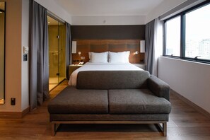 Een moderne hotelkamer met een groot bed, een slaapbank, een douchecabine met glazen wanden en uitzicht op de stad.