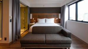 A modern hotel room with a large bed, a sofa, a glass shower area, and a city view.