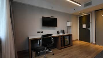 Quarto de hotel com secretária de madeira, televisão de ecrã plano na parede, um pequeno armário e uma porta à direita.