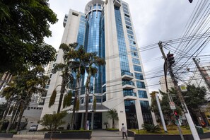 Un immeuble moderne de grande hauteur, doté d’une façade en verre, entouré de palmiers et bordé d’un passage piéton.