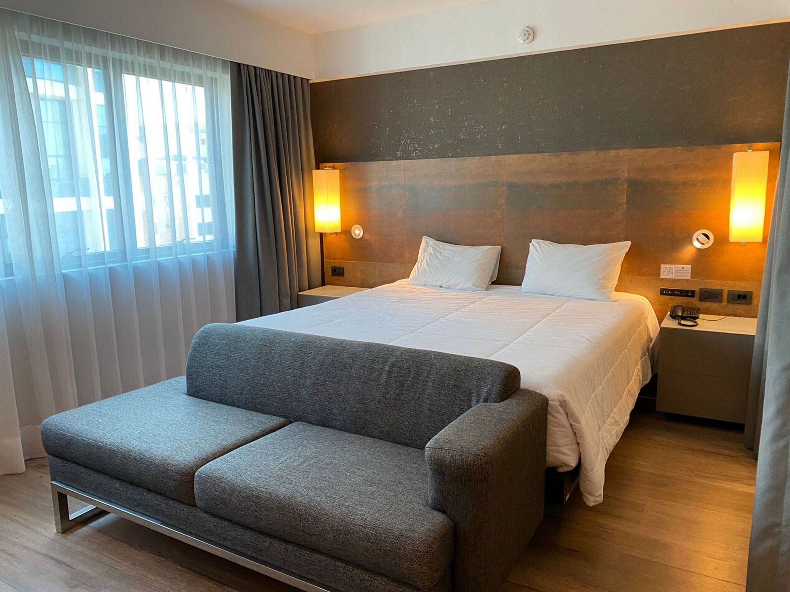 Una camera d'hotel con un letto, comodini, una poltrona e vista sull'esterno.