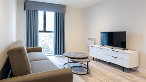 En moderne stue med en sofa, et rundt sofabord, et fjernsyn på et hvidt skab og et vindue med gardiner.