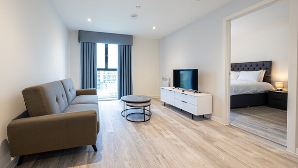 Et moderne hotelværelse med sofa, fjernsyn på et hvidt skab, seng med mørk sengegavl og udsigt over byen gennem store vinduer.
