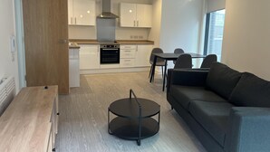 Eine moderne Küche mit weißen Schränken, einem eingebauten Ofen und einer Edelstahl-Dunstabzugshaube. Es gibt einen Essbereich mit Tisch und Stühlen sowie ein graues Sofa im Wohnbereich.