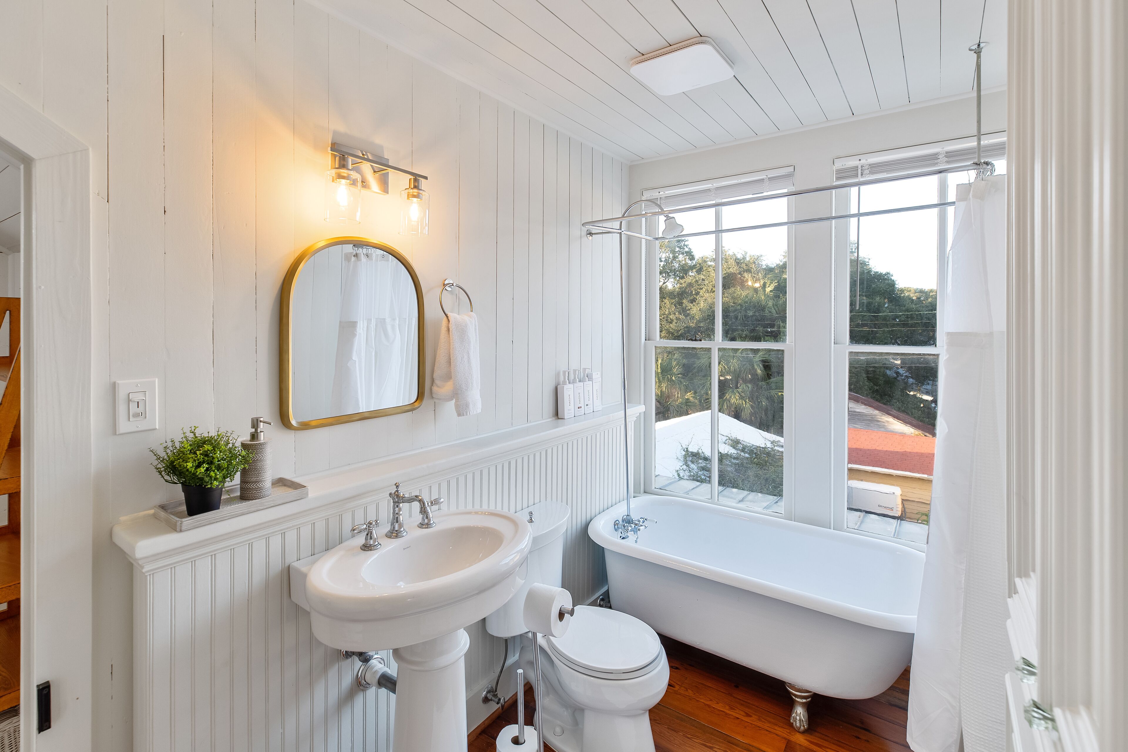 A bathroom with a white bathtub, a sink, and a toilet. There is a window with a view of trees and a red roof.
