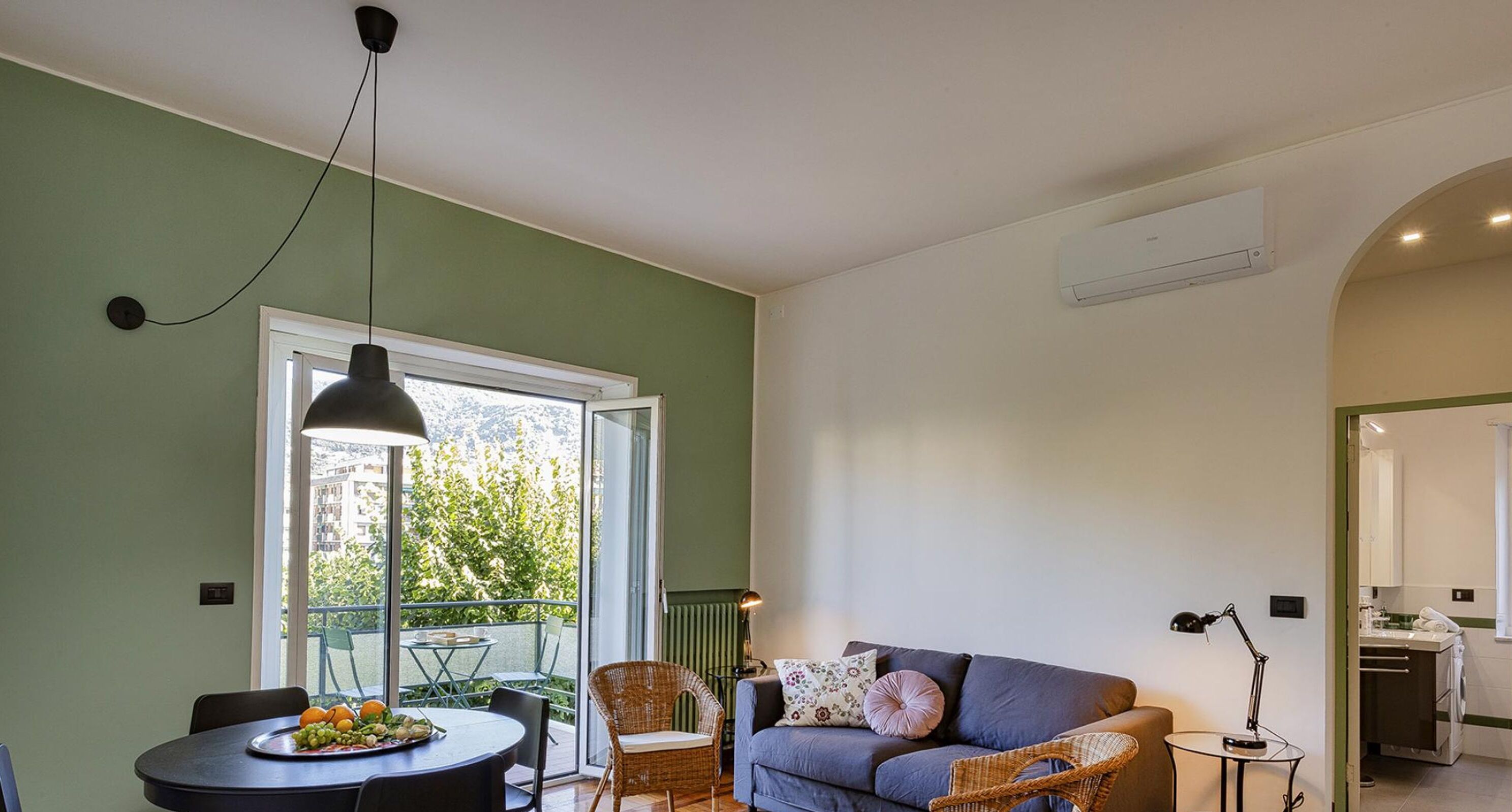 A room with a dining table, chairs, a sofa, a wicker chair, a lamp, and a window with a view of greenery.