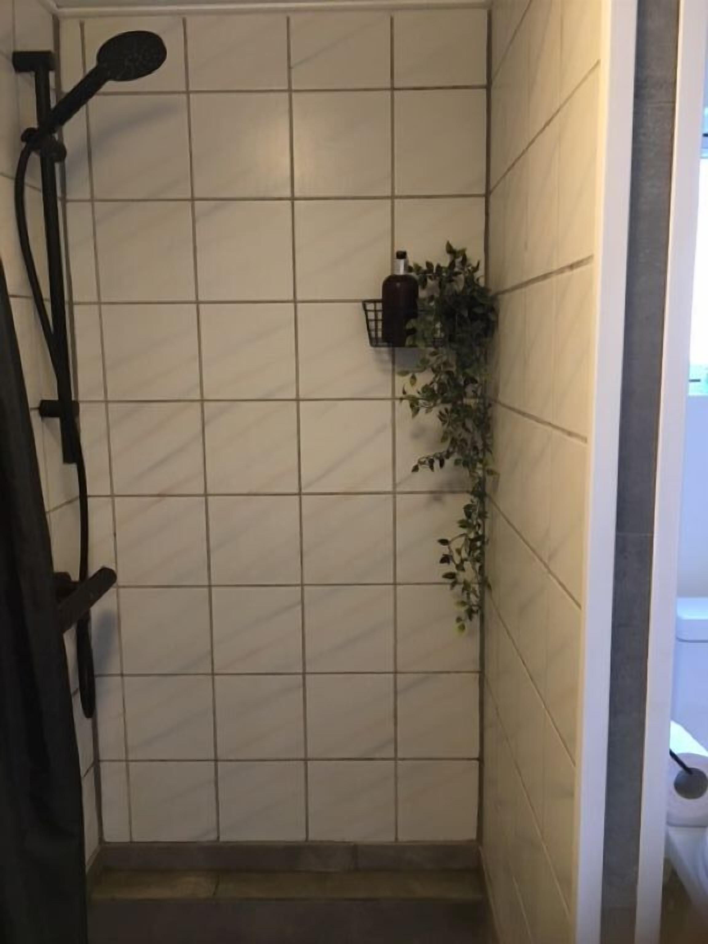 En duschhörna med kakel, en svart handduksställning, en liten hylla med en flaska och en krukväxt.