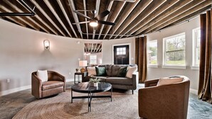 A spacious living room with a large sofa, two armchairs, a coffee table, and a woven rug. The room has exposed wooden beams on the ceiling and large windows with curtains. - HJ's Silos (Fayetteville)