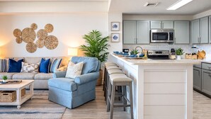A modern kitchen with grey cabinets, a central island, and a dining area with a blue sofa and a coffee table. - Gulf View Family Getaway, Amazing Pool, Short walk to Beach & Pier Park (Panama City Beach)