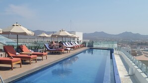 Ein Infinity-Pool auf dem Dach mit Liegestühlen und Sonnenschirmen, mit Blick auf Stadt und Berge.