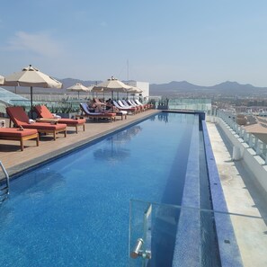 Piscina infinita en la azotea con sillones de descanso y sombrillas, con vistas a un paisaje urbano y montañas.