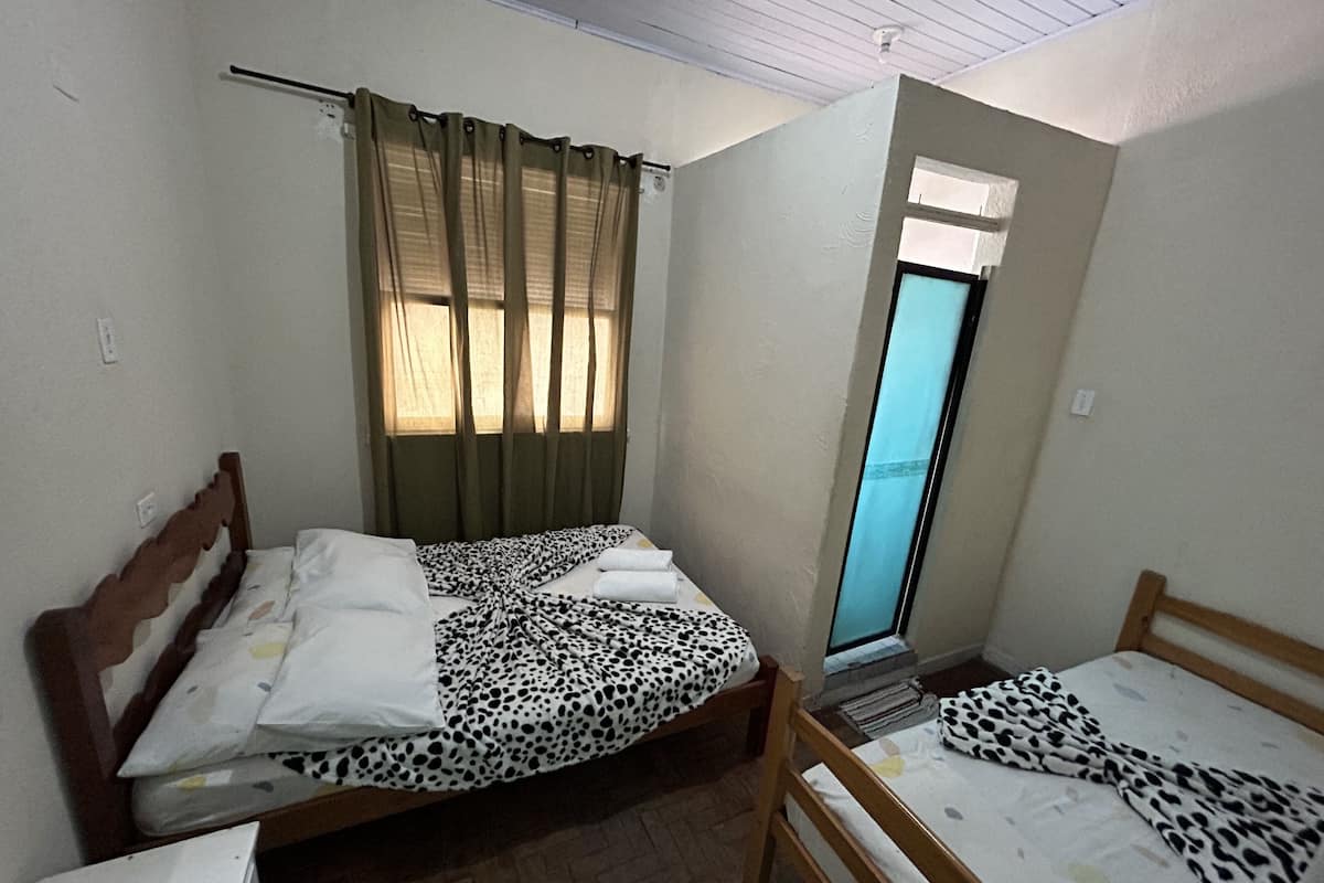 A small room with two beds, one with a leopard print bedspread and the other with a plain white bedspread. There is a window with curtains, a small table, and a mirror.