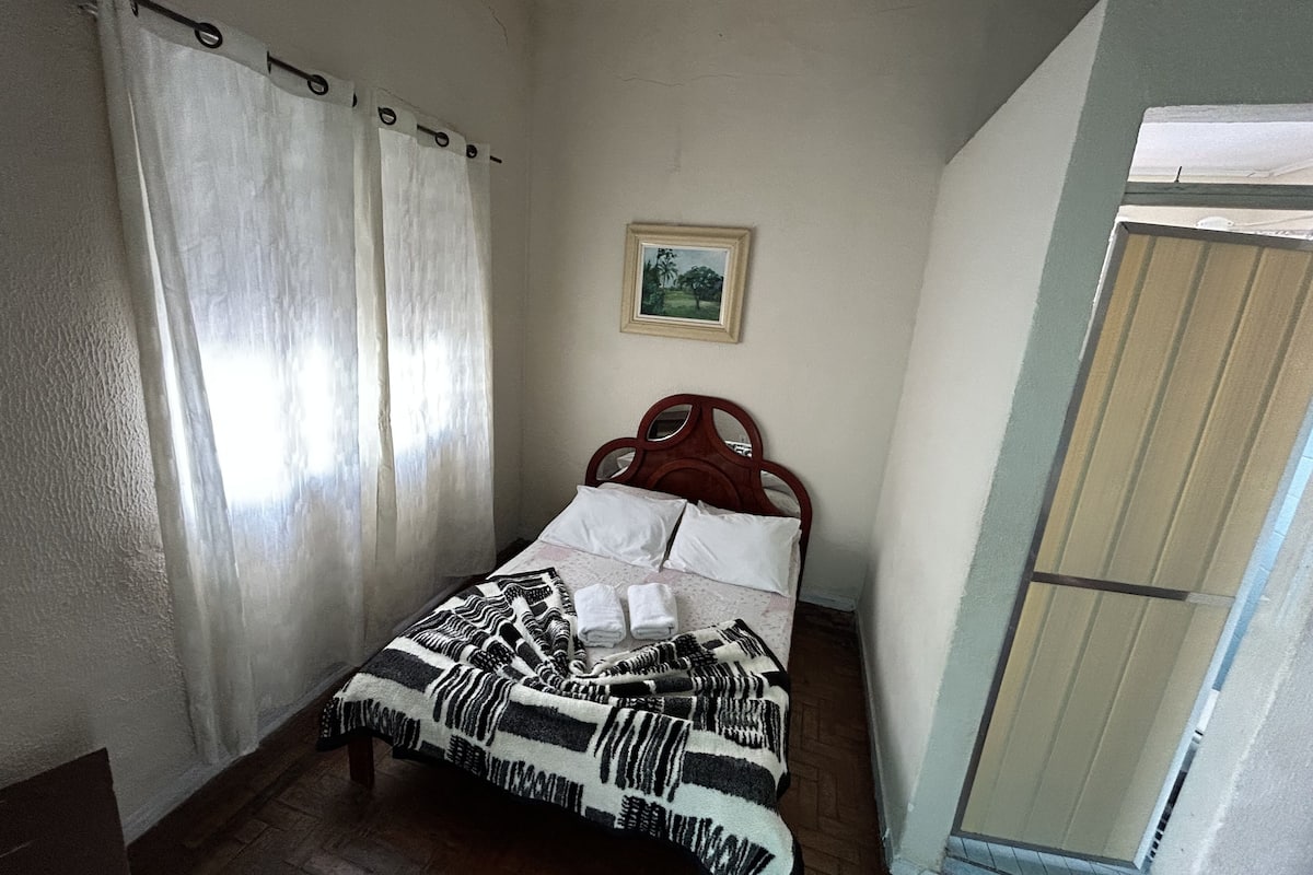 A bedroom with a bed, curtains, a framed picture on the wall, and a wooden door.