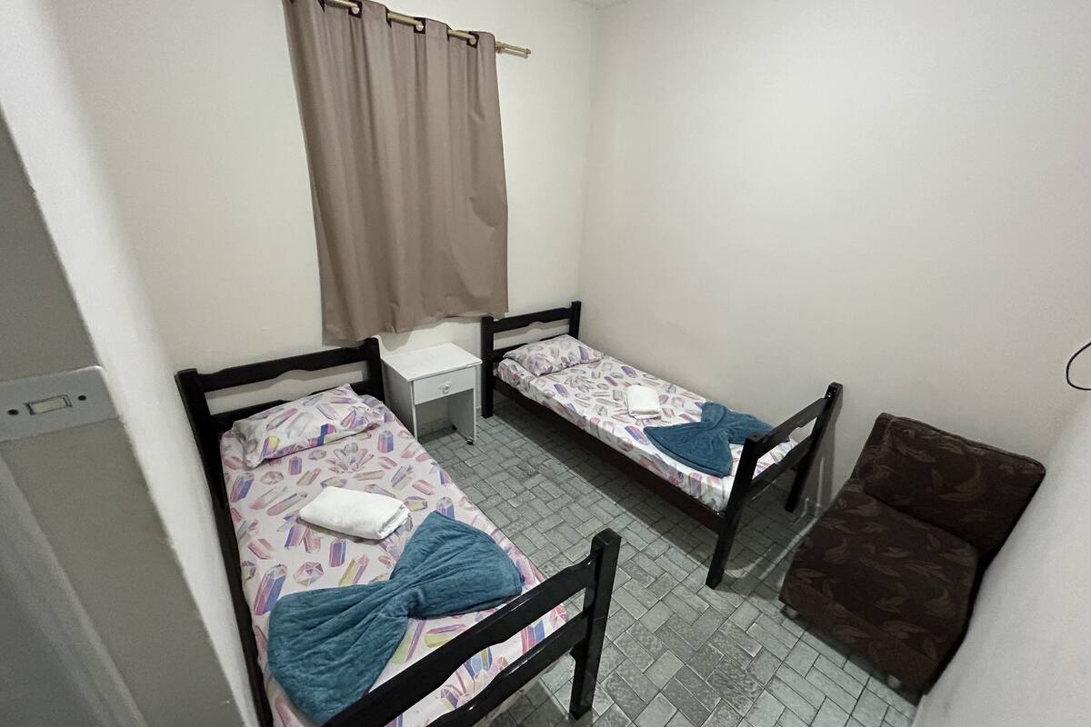 A dormitory room with two single beds, a small bedside table, a chair, and a window with curtains.