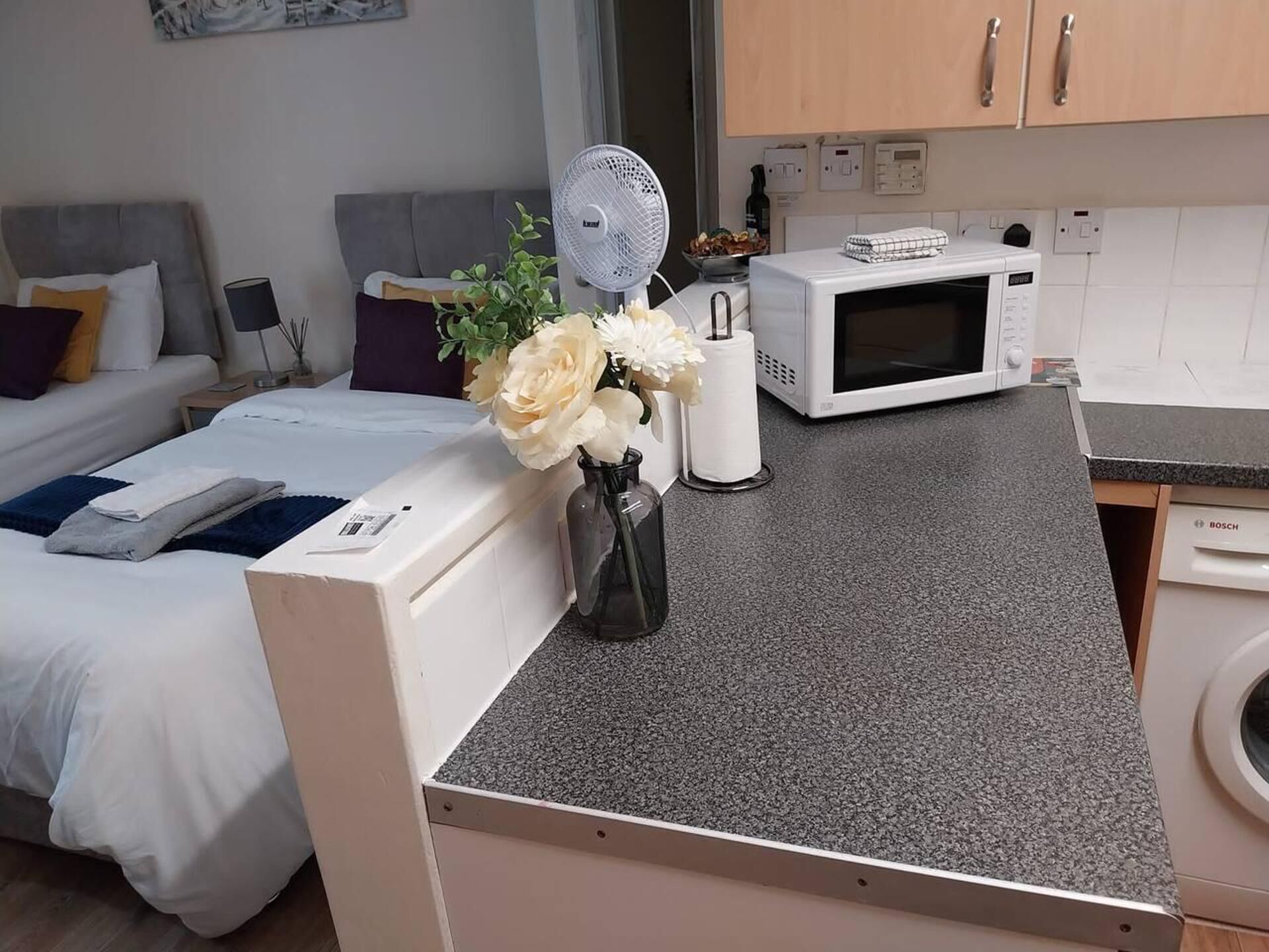 Ein kompaktes Hotelzimmer mit Bett, kleinem Schreibtisch, Mikrowelle, Waschmaschine und Ventilator.