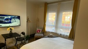 Quarto de hotel com cama, escrivaninha com televisão, cadeira e janela com cortinas.