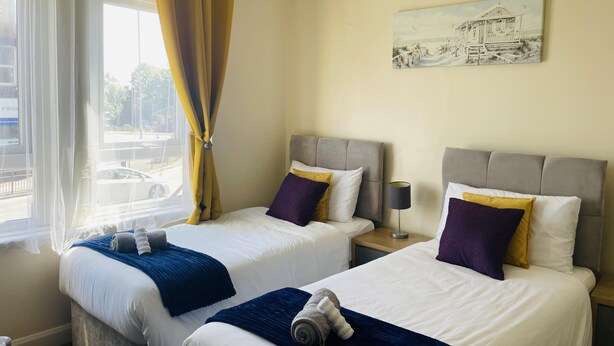 Quarto de hotel com duas camas, uma janela com cortinas, um quadro na parede e uma luminária na mesinha de cabeceira.