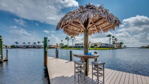A wooden dock with a thatched umbrella table and two stools, overlooking calm water with buildings and palm trees in the background. - Beautiful views, watch the dolphins and tarpon swim by on the  lanai or dock! (Cape Coral)