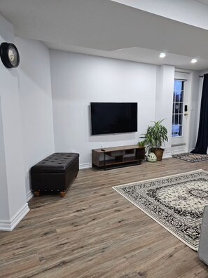 A living room with a flat-screen TV, a dark leather ottoman, a wooden entertainment unit, a patterned rug, and a potted plant.