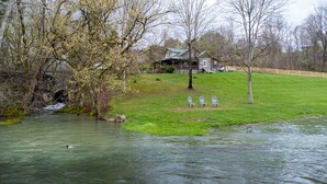 A riverside view with a wooden cabin, a grassy area with white chairs, and a duck swimming in the water. - Brown Trout (Bristol)
