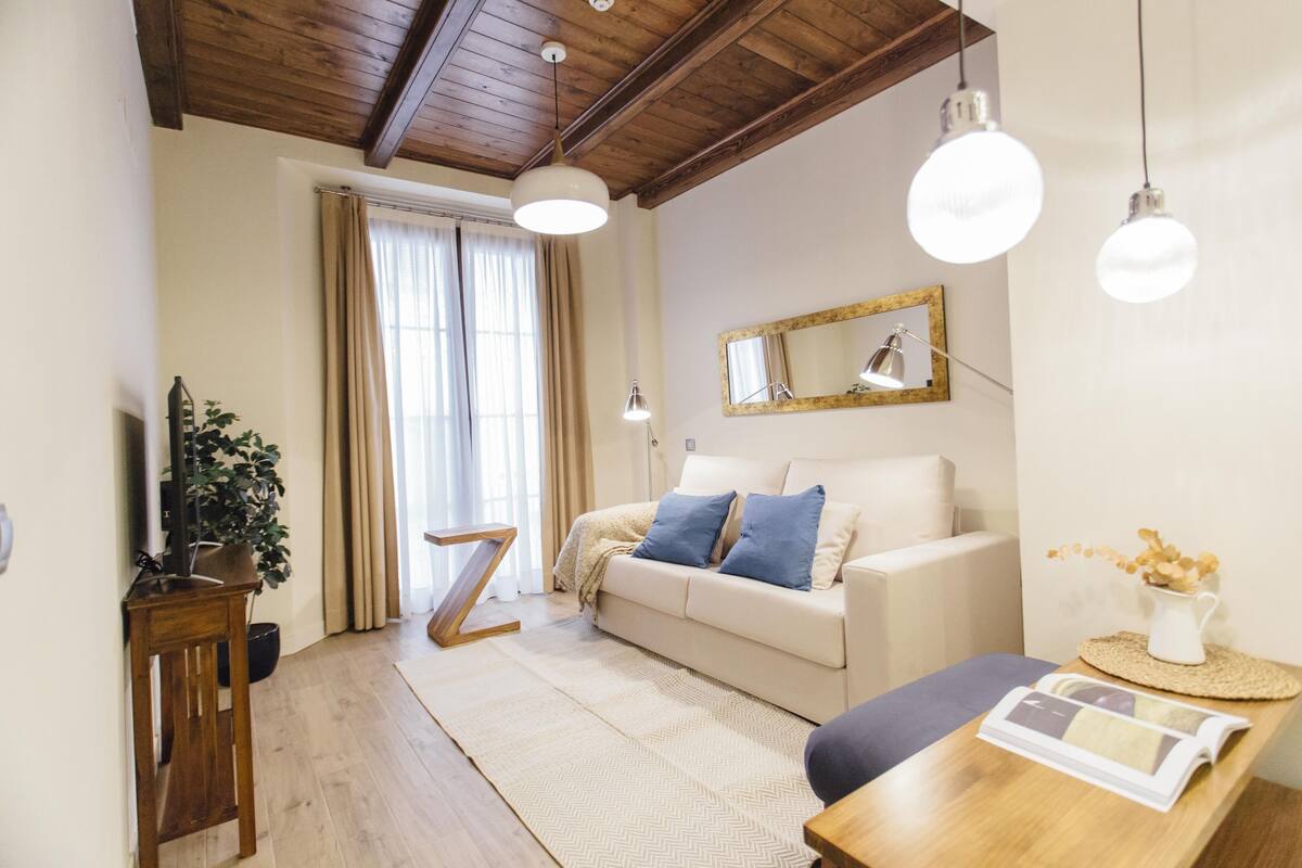 A cozy living room with a sofa, a coffee table, and a wooden side table. There is a large window with curtains, a plant, and a mirror on the wall.