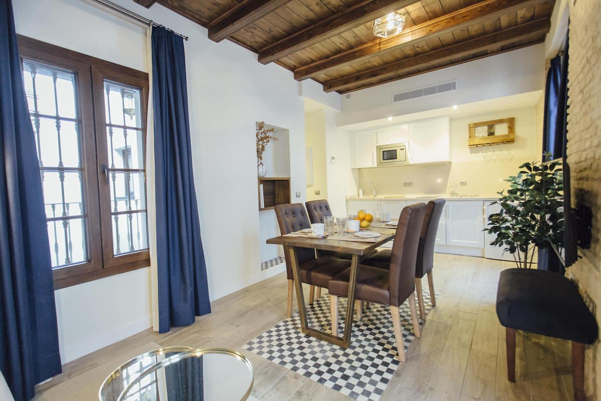 A dining area with a checkered floor, wooden table, and chairs. There is a kitchen in the background with white cabinets and a microwave.