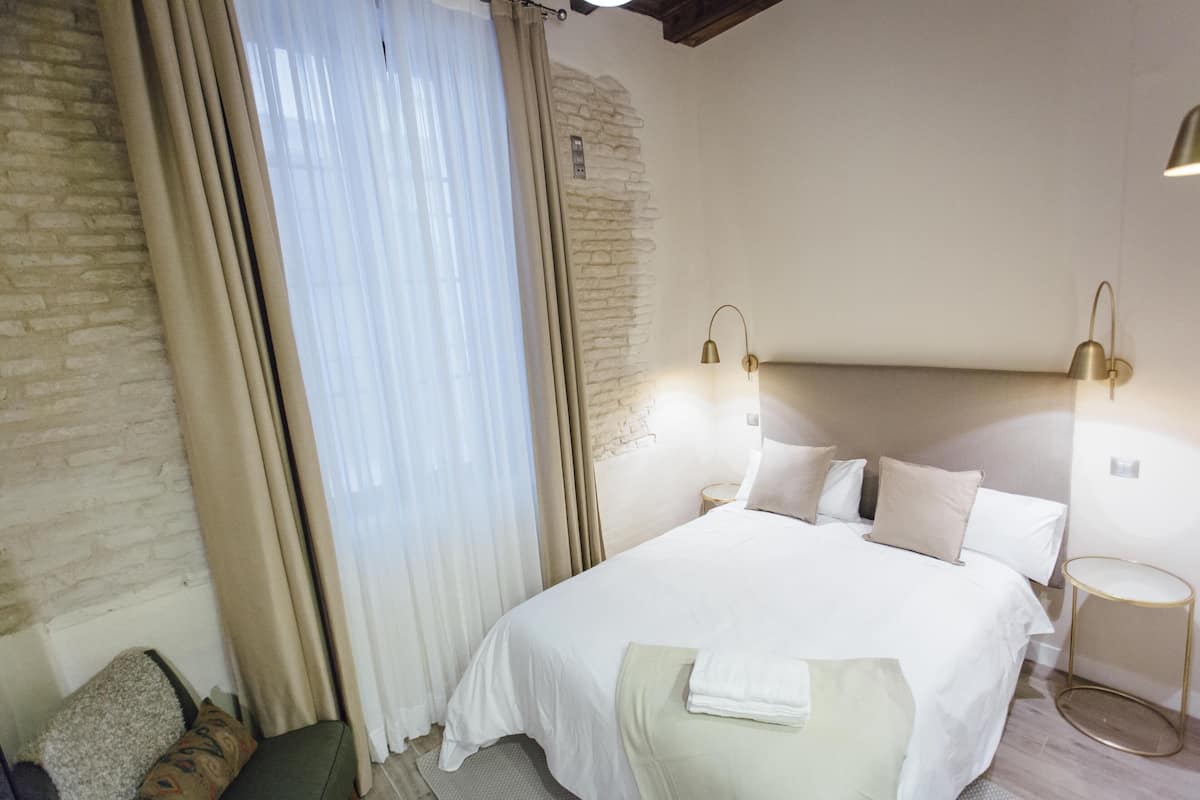 A neatly made bed with white linens, a bedside table with a lamp, and a window with sheer curtains.