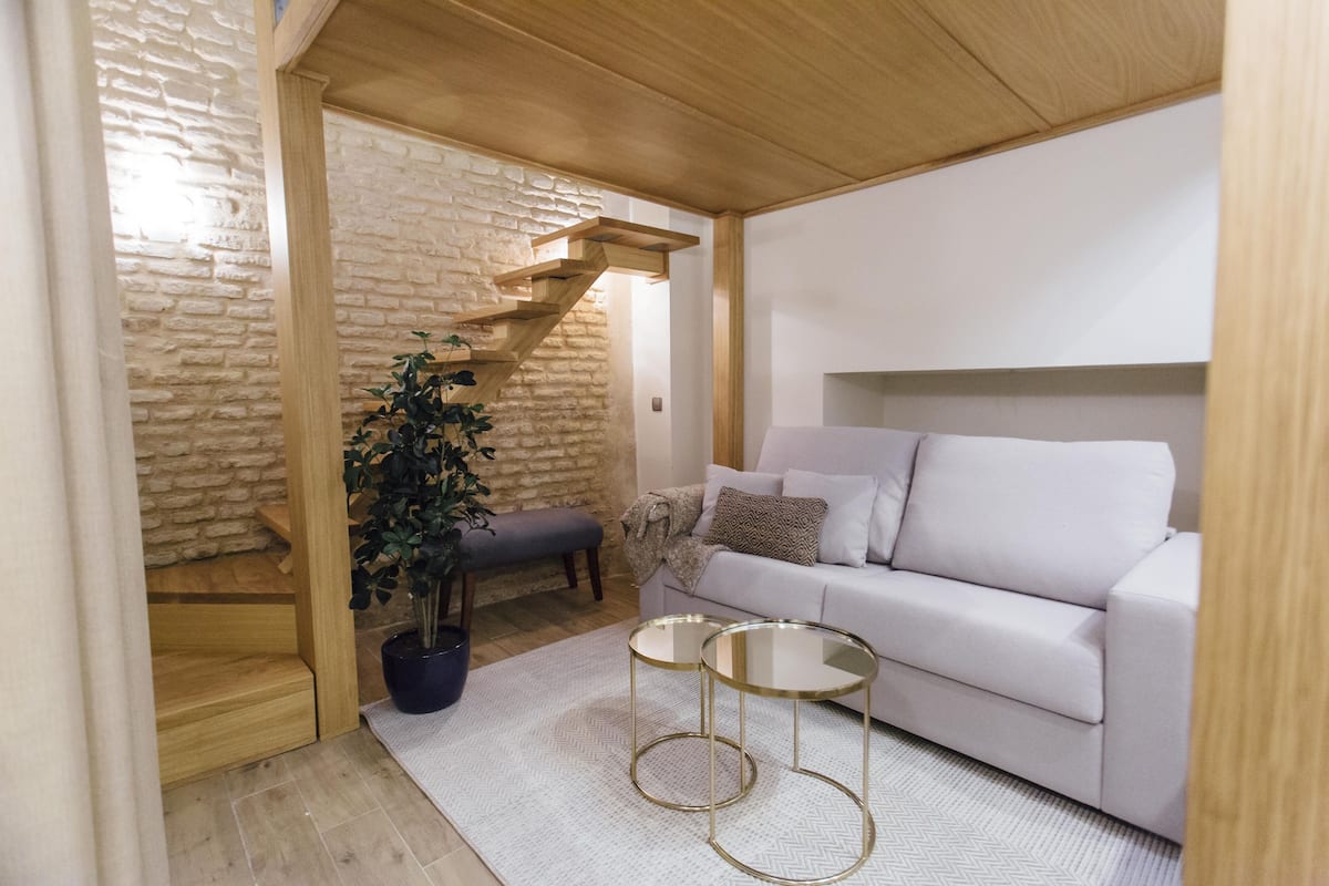 A modern living room with a white sofa, two glass-top side tables, a potted plant, and a wooden staircase.