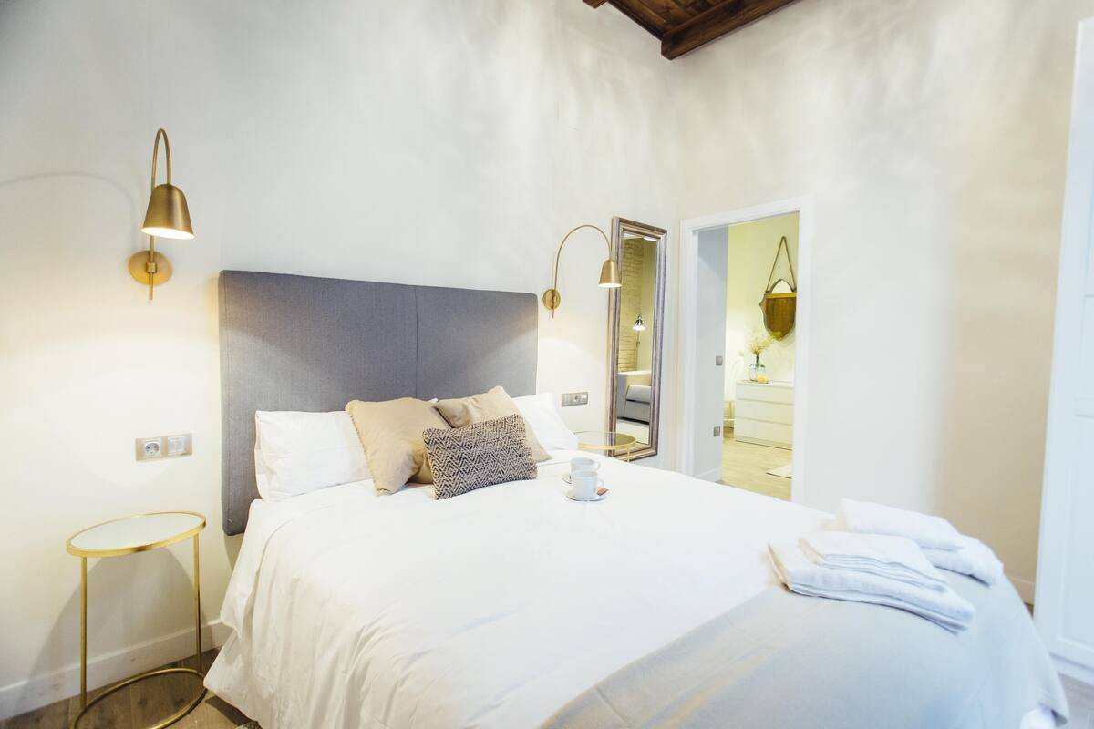 A neatly made bed with white linens, a grey headboard, and a bedside table with a lamp.