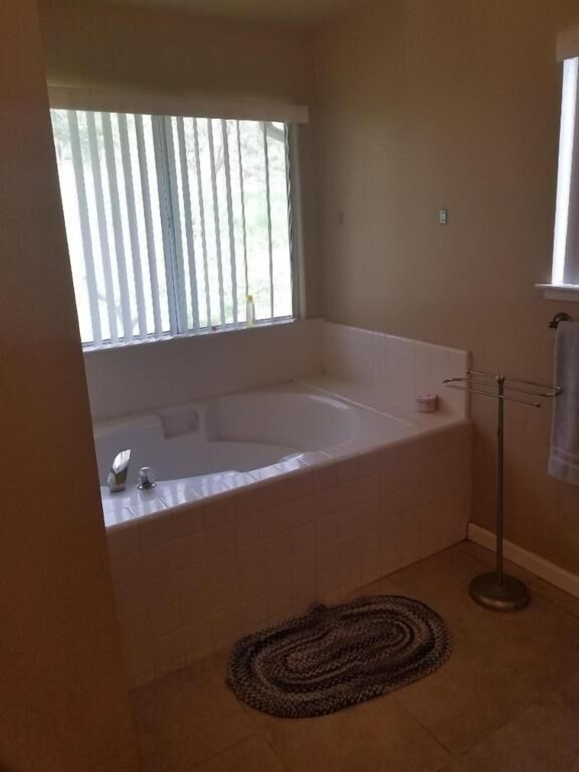 A bathroom with a bathtub, a window with blinds, a towel rack, and a rug.