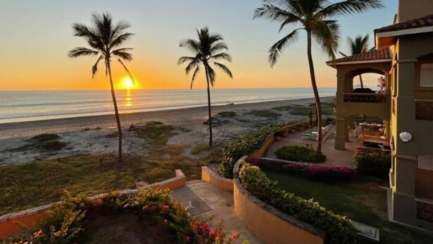Una propiedad frente a la playa con vista al atardecer sobre el océano, palmeras y un jardín paisajista.