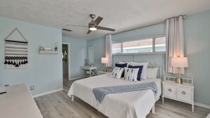 Ein Schlafzimmer mit einem Bett, Nachttischen, einem Deckenventilator, einem Fenster mit Vorhängen und einer Wanddekoration.