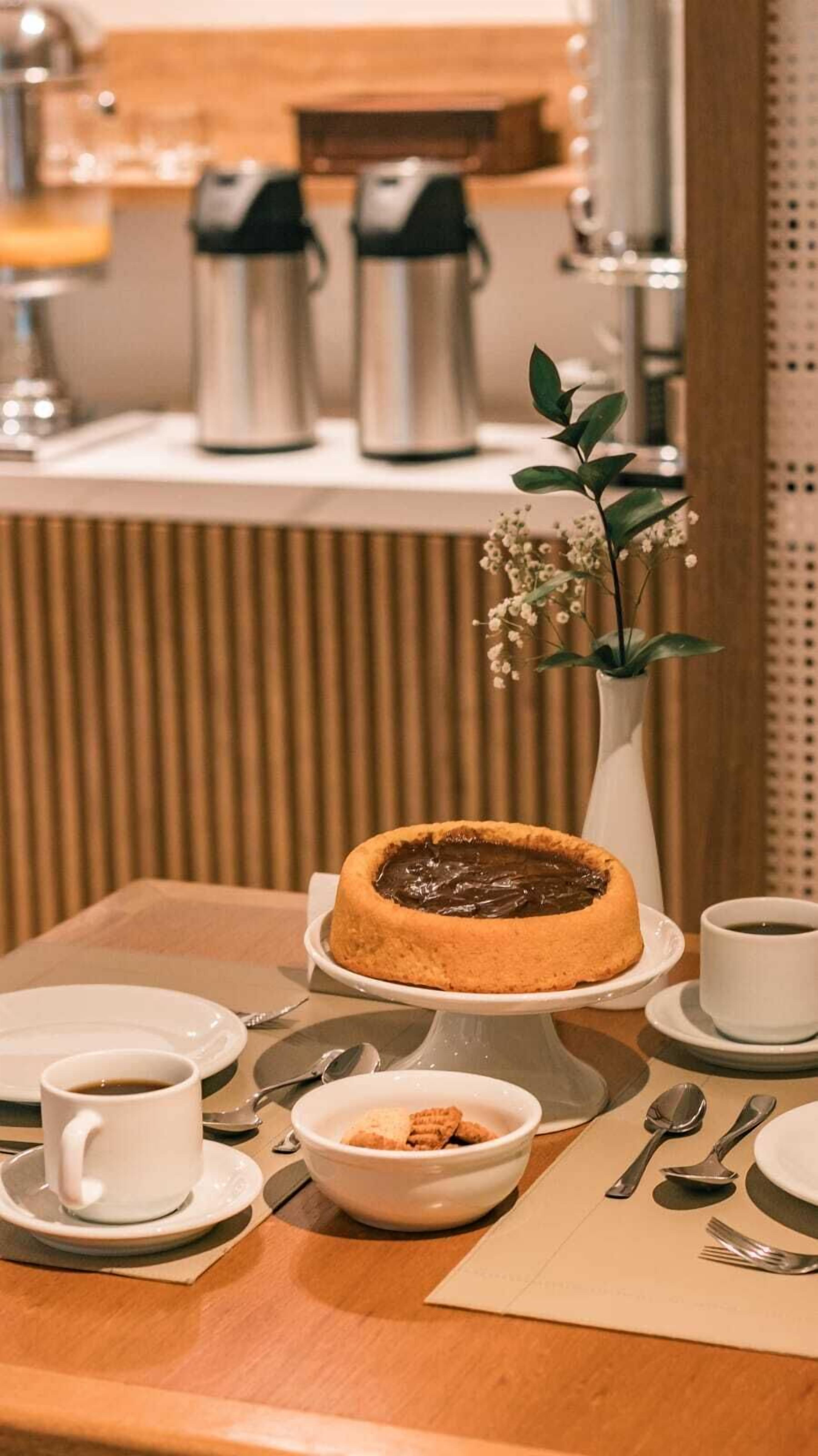 Una mesa de postres con un pastel de chocolate, tazas de café y un bol pequeño de galletas.