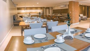 A modern dining area with tables set for breakfast, a brick pillar, and a seating area with sofas and a coffee station. - Village du Lac Hotel (Pomerode)