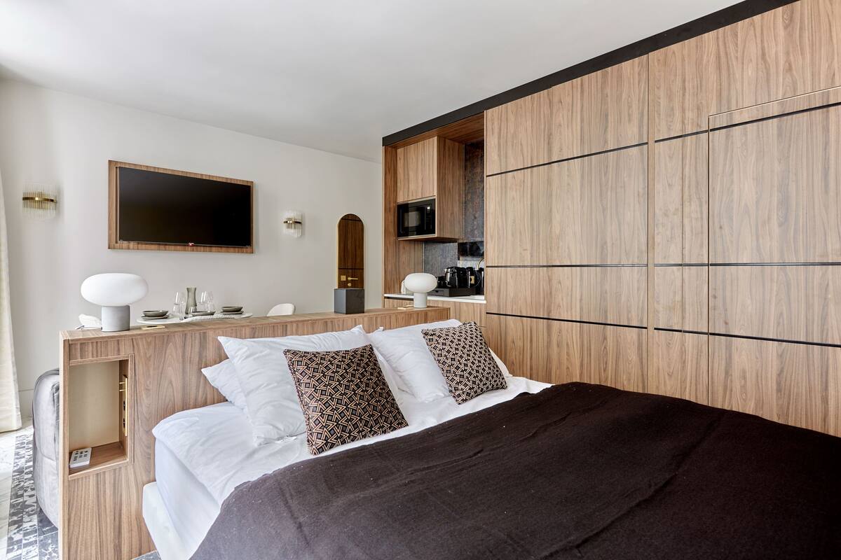 A modern hotel room with a large bed, a wooden wardrobe, a built-in kitchen area, and a flat-screen TV mounted on the wall.