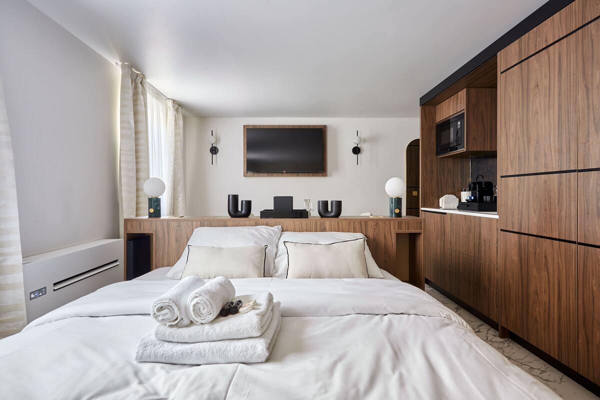 A hotel room with a large bed, wooden wardrobe, flat-screen TV, and a small kitchen area with a microwave.