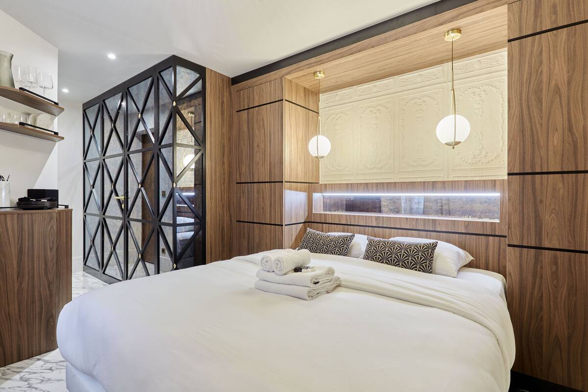 A modern hotel room with a large bed, wooden paneling, a geometric wall design, and a built-in light fixture.
