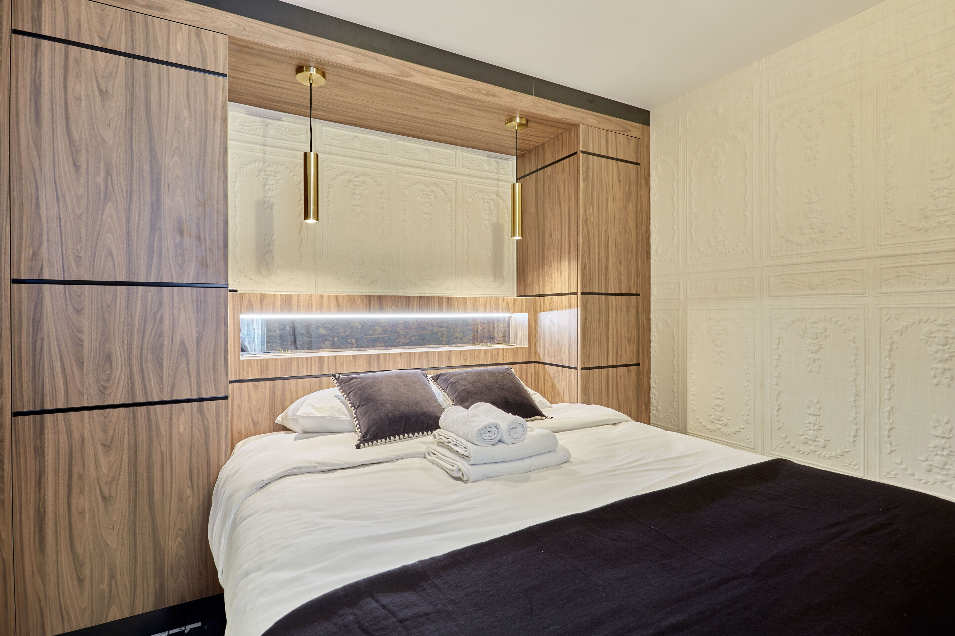 A modern bedroom with a wooden headboard, a large window, and a neatly made bed with towels.