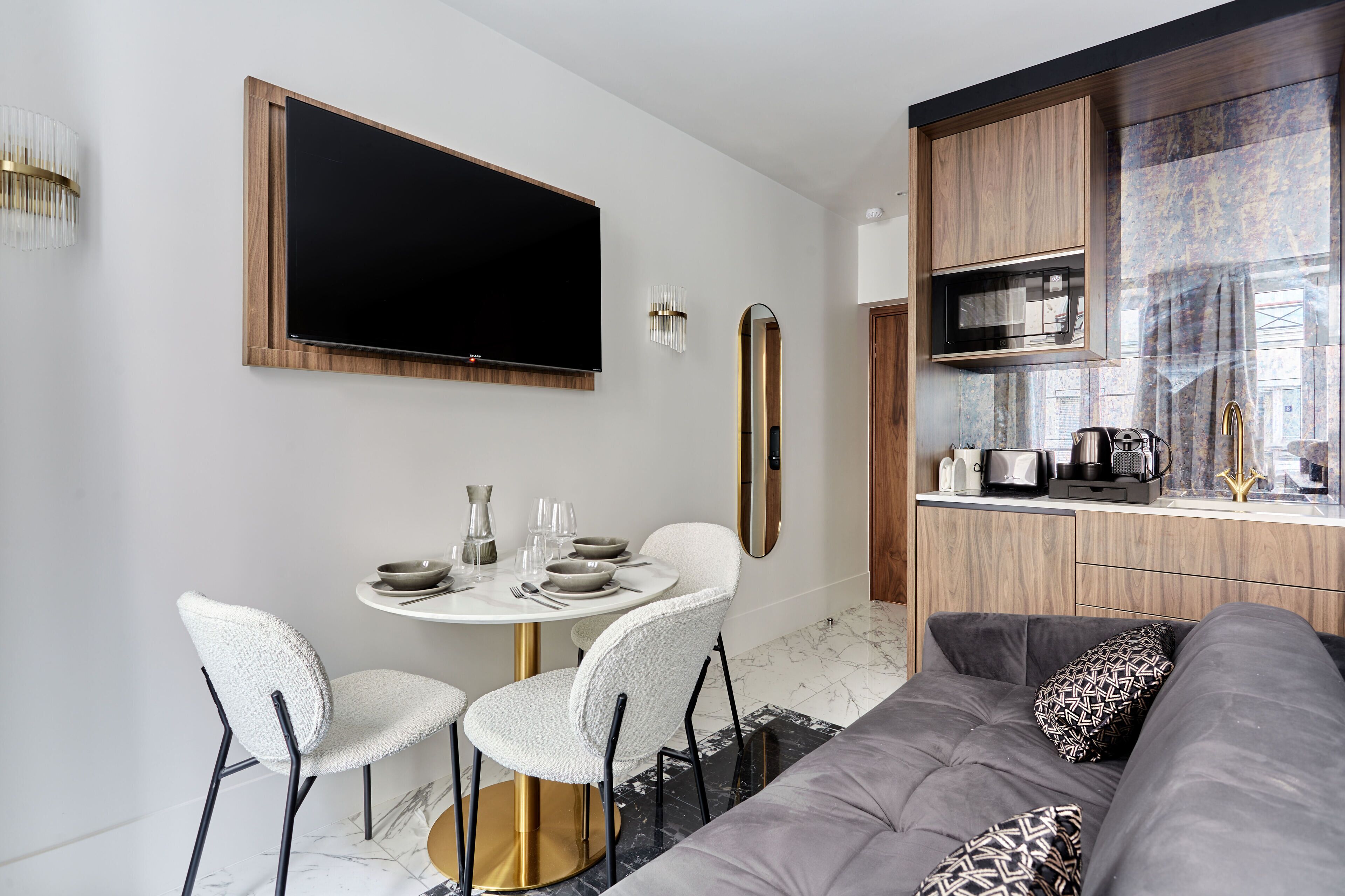 A modern living room with a grey sofa, a round dining table with white chairs, a flat-screen TV mounted on the wall, and a wooden kitchen area with a microwave.
