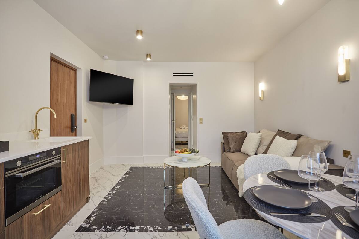 A modern living room with a sofa, a dining table set for two, a kitchen area with a built-in oven, and a flat-screen TV mounted on the wall.