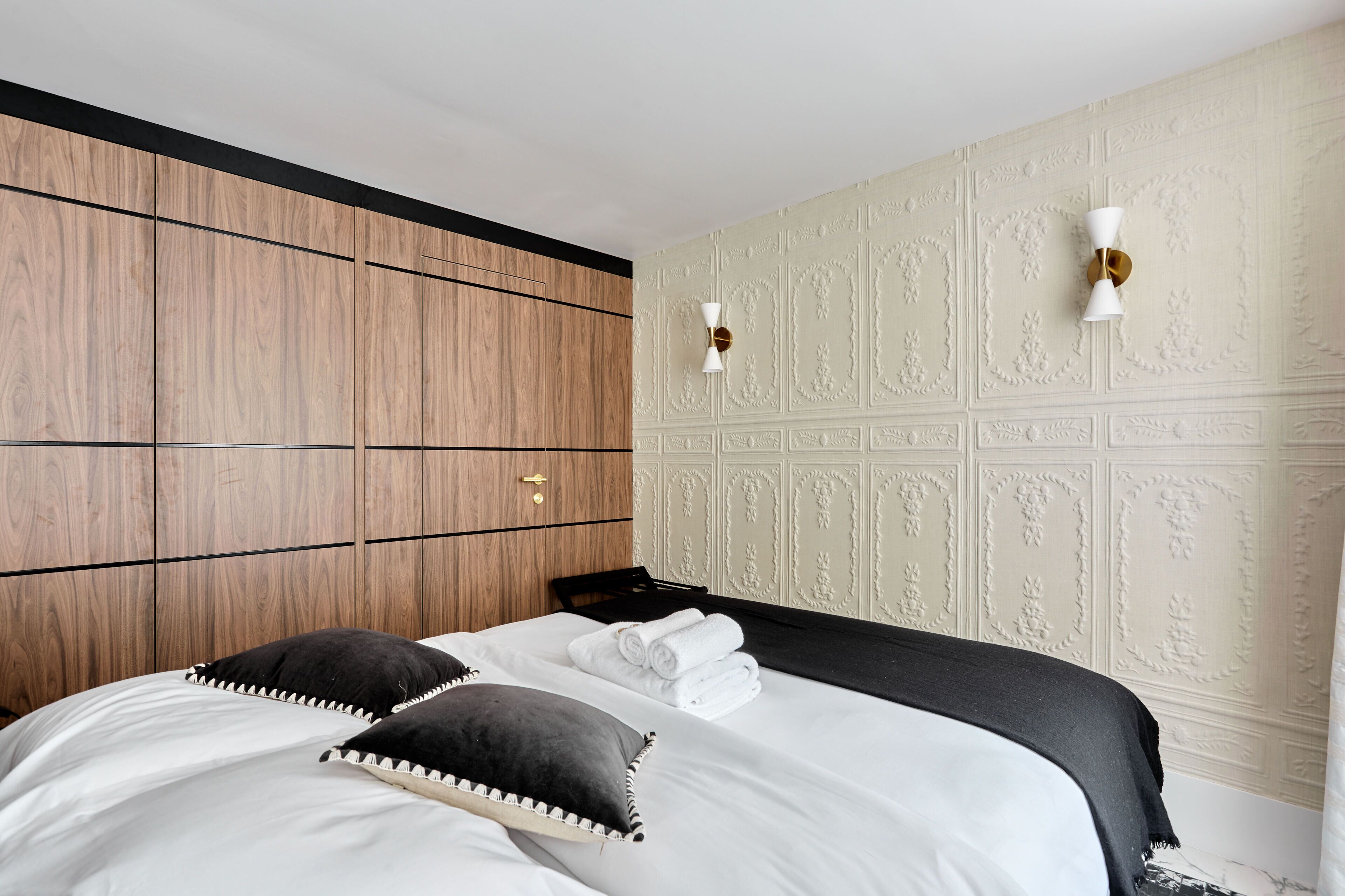 A hotel room with a wooden headboard, a bed with white linens and black pillows, a black throw blanket, and decorative wall sconces.