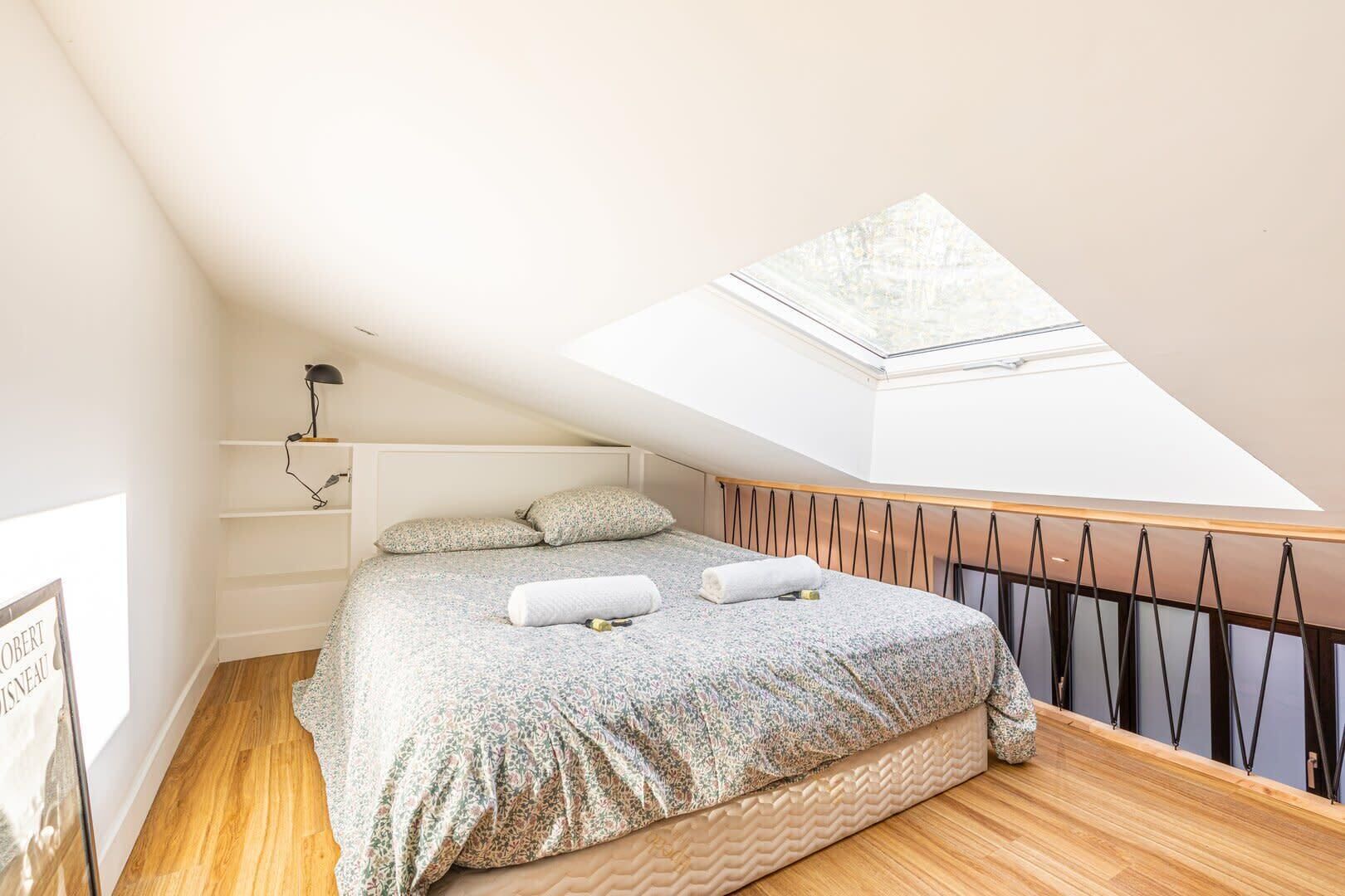 A bedroom with a skylight, a bed with floral bedding, a bedside table, and a wall-mounted shelf.