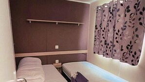 A compact room with two beds, a small table, and a window with curtains. - Spacious chalet with furnished terrace (Fréjus)